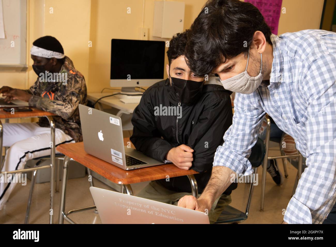 Educación Maestro masculino de la High School que se inclina para mostrar a estudiante masculino algo en la computadora portátil, ambos usando máscaras faciales para proteger contra Covid-19 Foto de stock
