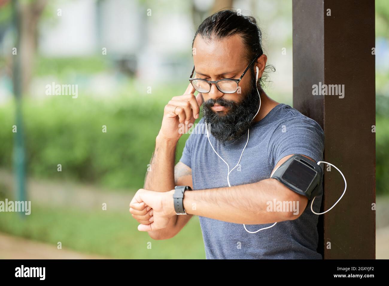 Hombre indio barbudo joven serio en anteojos de pie al aire libre y ajustar  los auriculares mientras escucha mensajes de audio en smartwatch Fotografía  de stock - Alamy