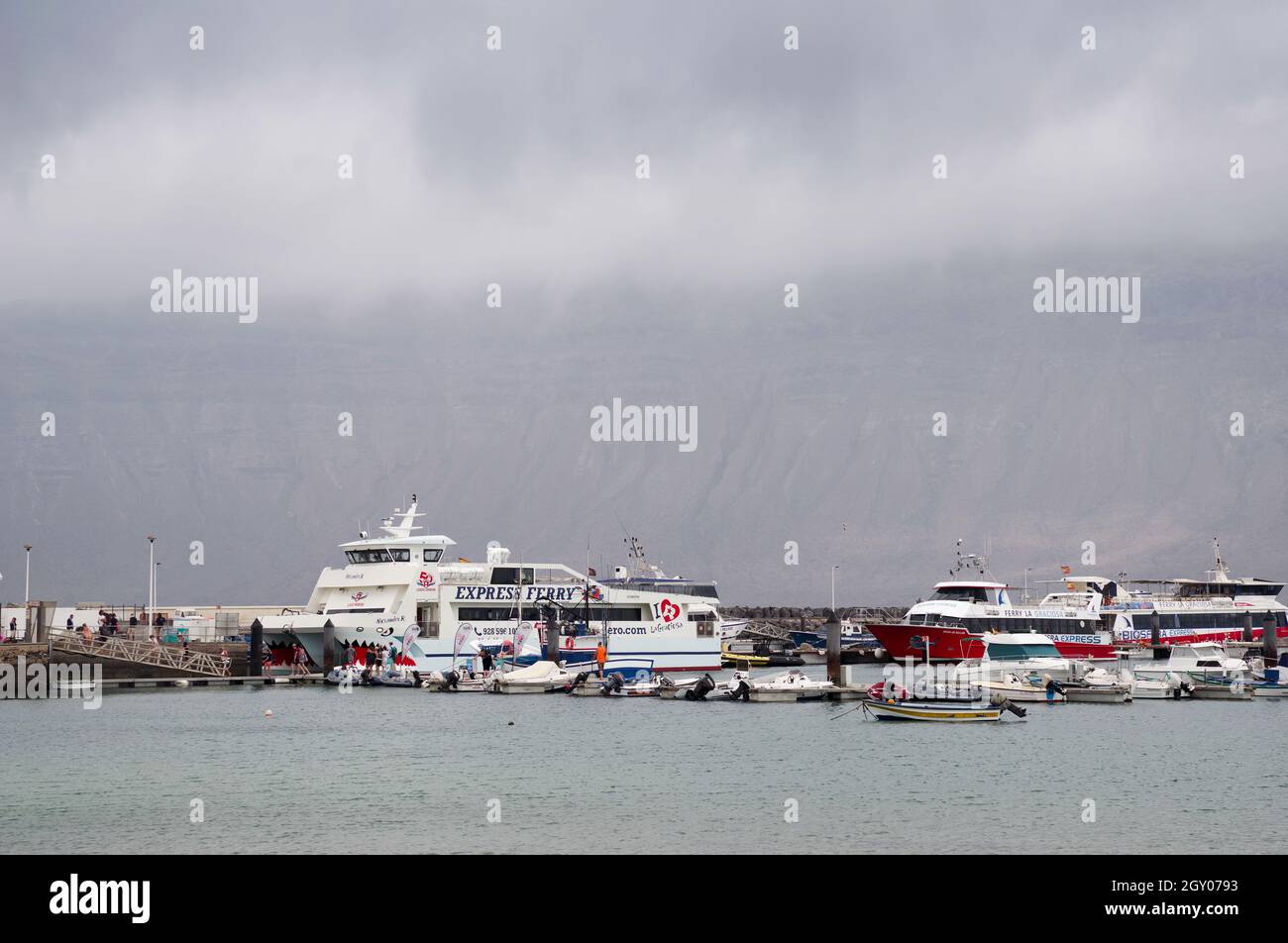 Barcos y ferry amarrados en la isla de La Graciosa, nubes que cubren los acantilados de Lanzarote en la distancia Foto de stock