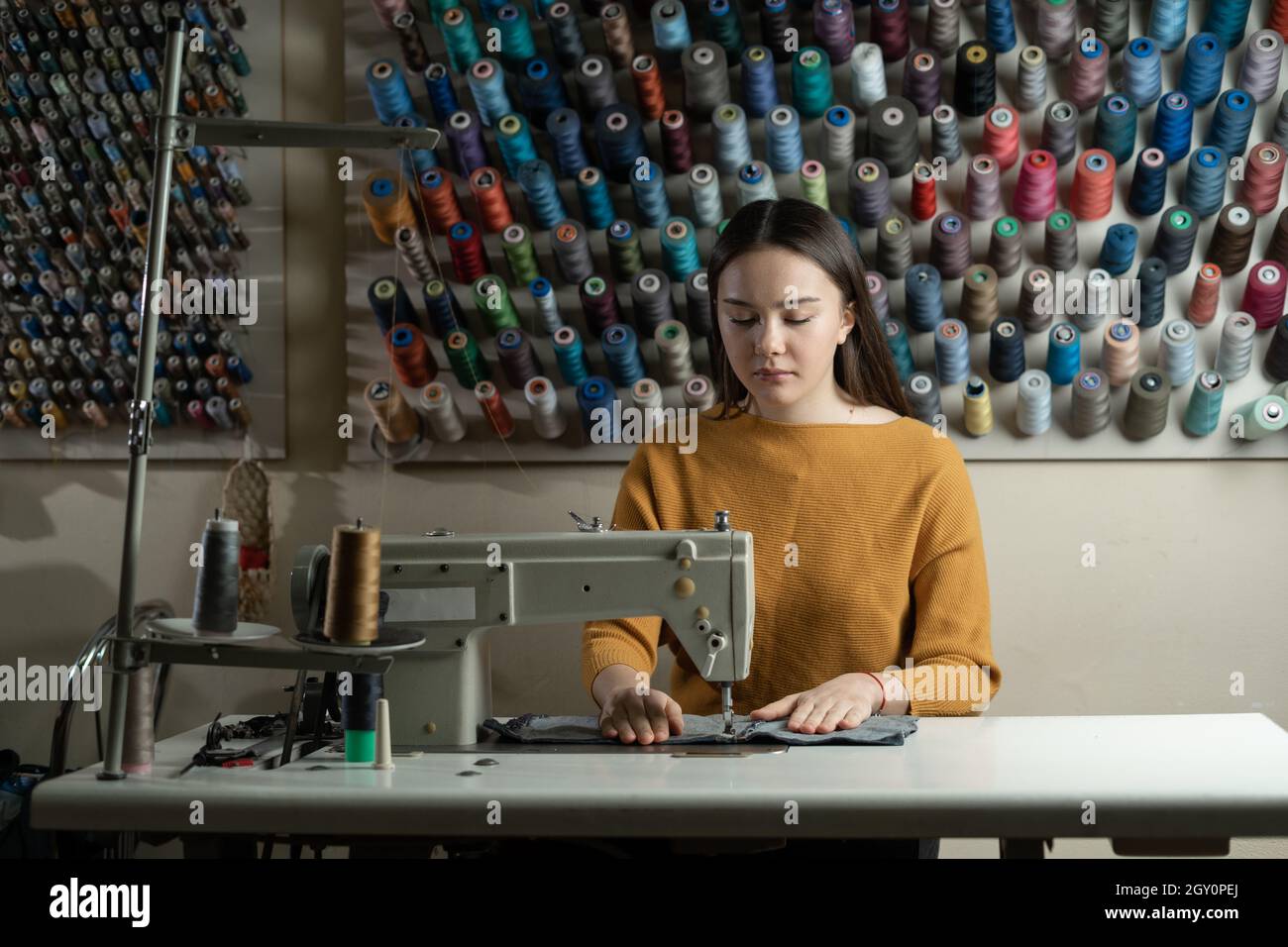 Una mujer trabaja en una máquina de coser en un taller. Ella se sienta en  la mesa y soda ropa de algodón. Fondo de bobinas de hilo de colores  diferentes Fotografía de