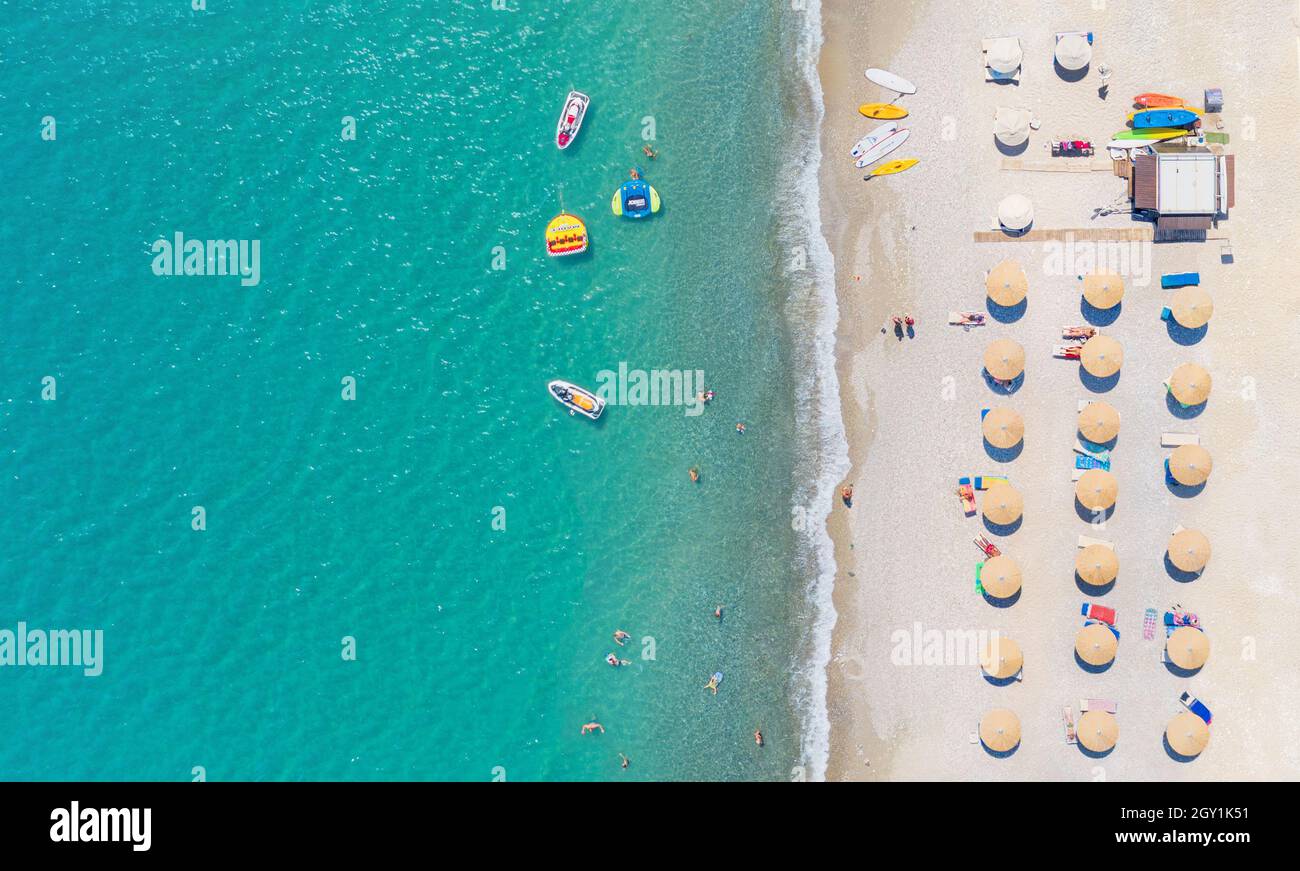 Playa de arena con tumbonas, sombrillas y equipos de deportes acuáticos, gente nadando y tomando el sol. Panorama aéreo directamente sobre Foto de stock