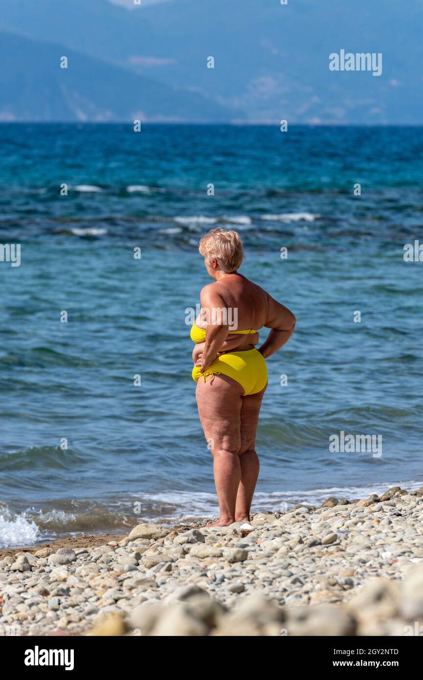 una mujer de mediana edad con sobrepeso que llevaba un bikini amarillo de  pie en la playa de la orilla del mar con las manos en las caderas mirando  hacia el mar.