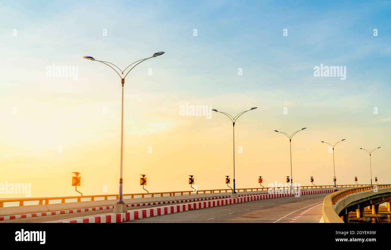 Curva carretera de hormigón con curva y señal de tráfico a pie al lado del mar al atardecer. La energía del panel solar en la señal de tráfico de la curva amarilla. Viaje por carretera Foto de stock