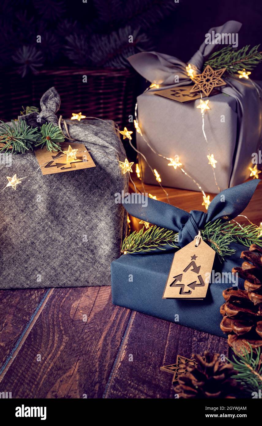 Tres cajas de regalo ecológicas estilo furoshiki envueltas sobre fondo oscuro con luces y etiquetas de regalo. Cero residuos Navidad Reciclaje, reutilización, sostenible l Foto de stock