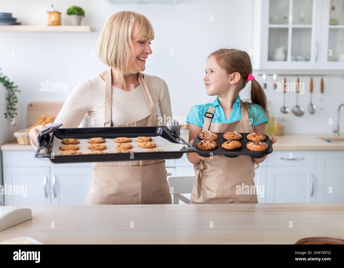 Aprender a cocinar. Retrato de abuela madura sonriente y nieta que muestra sabrosas galletas recién horneadas y magdalenas en la bandeja mientras está de pie Foto de stock