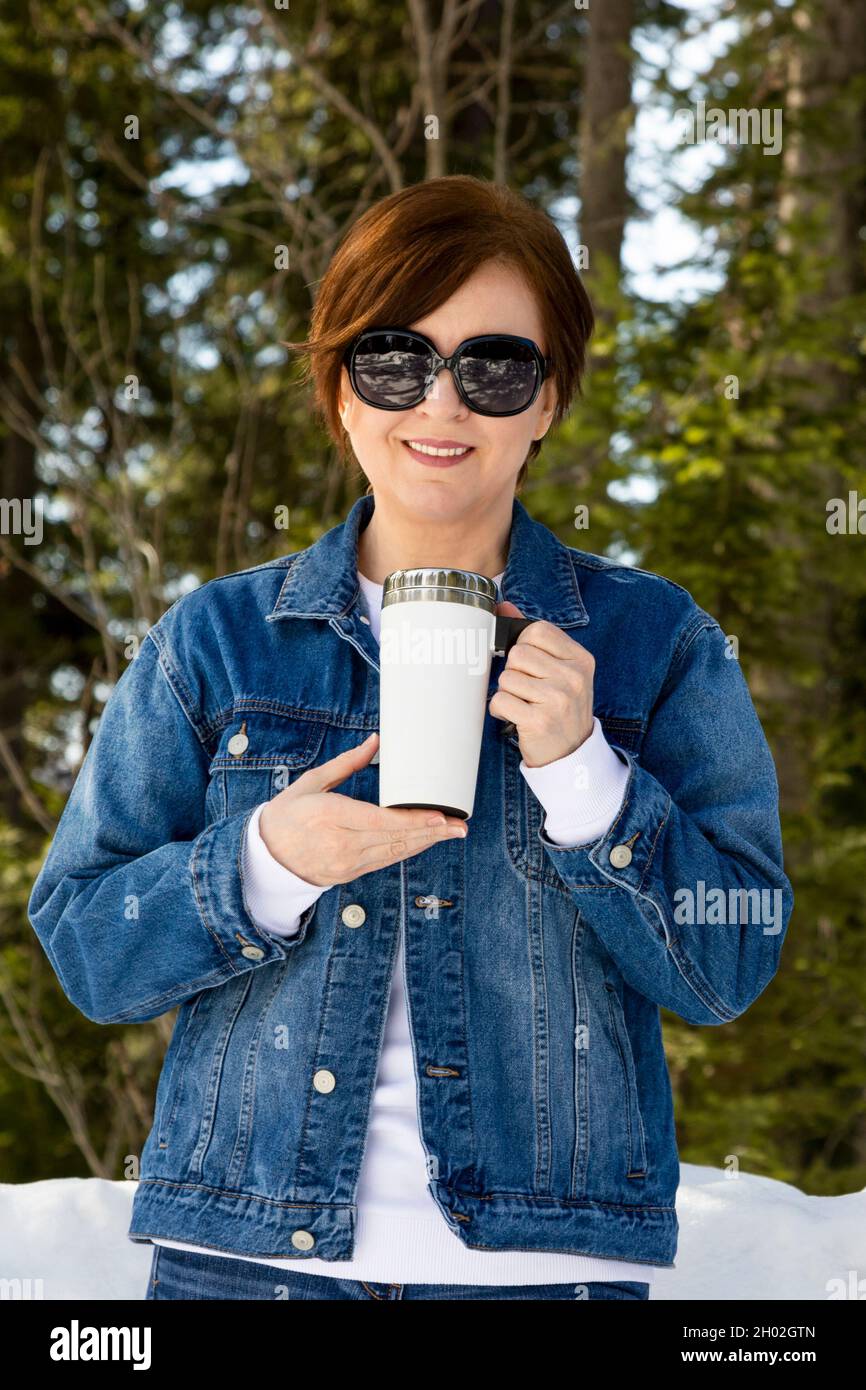 Se burló de una mujer que llevaba gafas de sol sosteniendo una taza blanca  de viaje junto a un bosque nevado. Taza de café de viaje vacía con plantilla  de mango, maqueta
