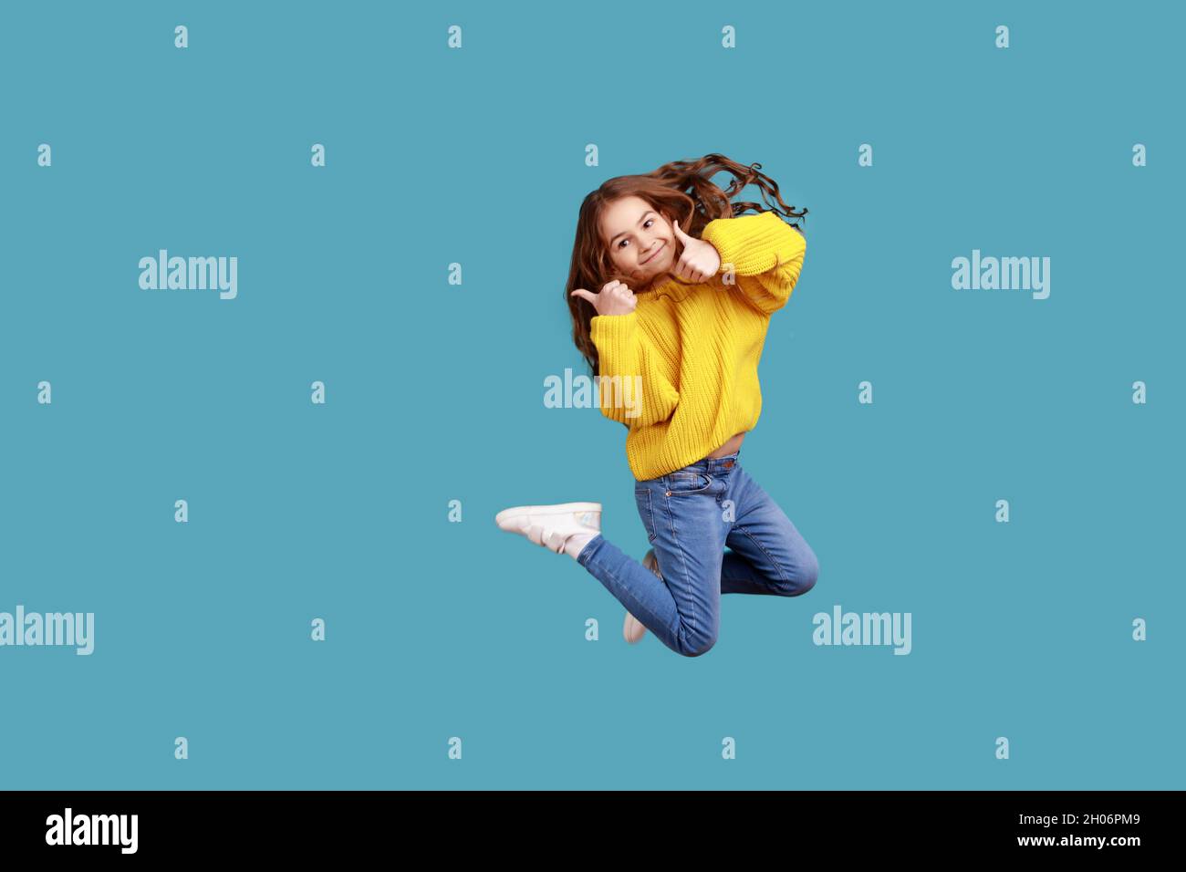 Retrato completo de feliz encantadora niña saltando alto y mostrando el pulgar hasta la cámara, usando un jersey amarillo estilo informal. Estudio en interior grabado aislado sobre fondo azul. Foto de stock