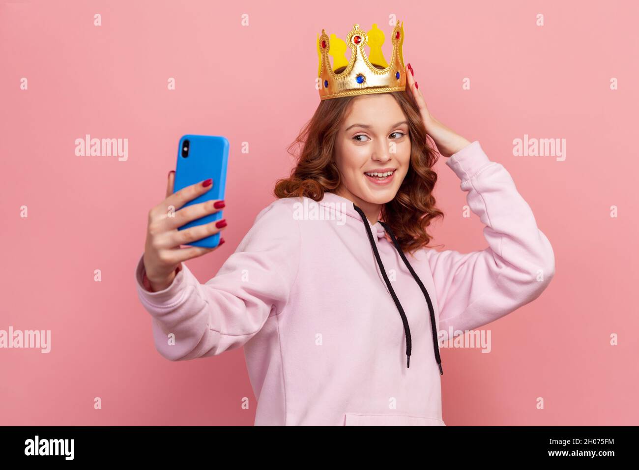 Retrato de una chica adolescente sonriente de pelo rizado con capucha y corona dorada posando en la cámara del smartphone, tomando selfie. Estudio en interior grabado aislado sobre fondo rosa Foto de stock