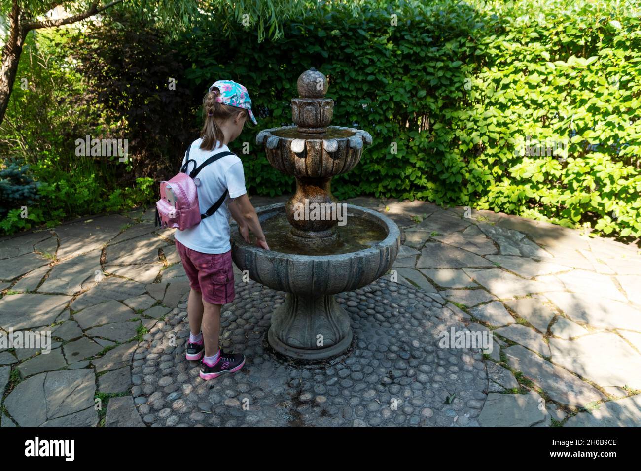 Una chica en una gorra y con una mochila llita su mano en un tazón de fuente en miniatura de pie a la sombra de los árboles en un día caluroso de verano. Foto de stock