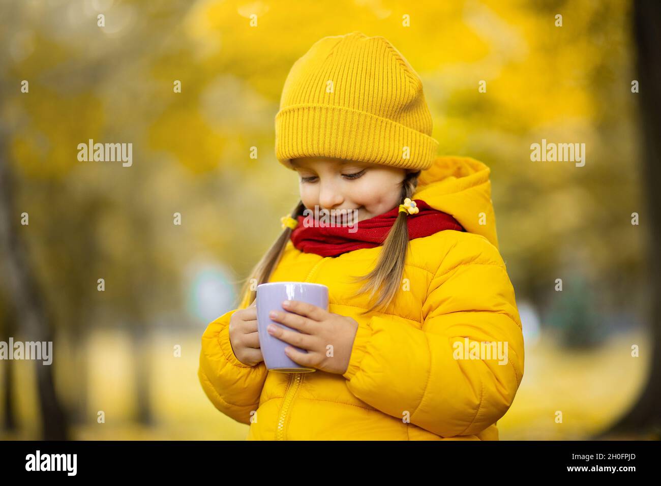 Foto de otoño cálido. Foto de cerca de una linda niña pequeña con abrigo y  gorra amarillo brillante, sosteniendo una taza de chocolate caliente o té  en el parque dorado de otoño