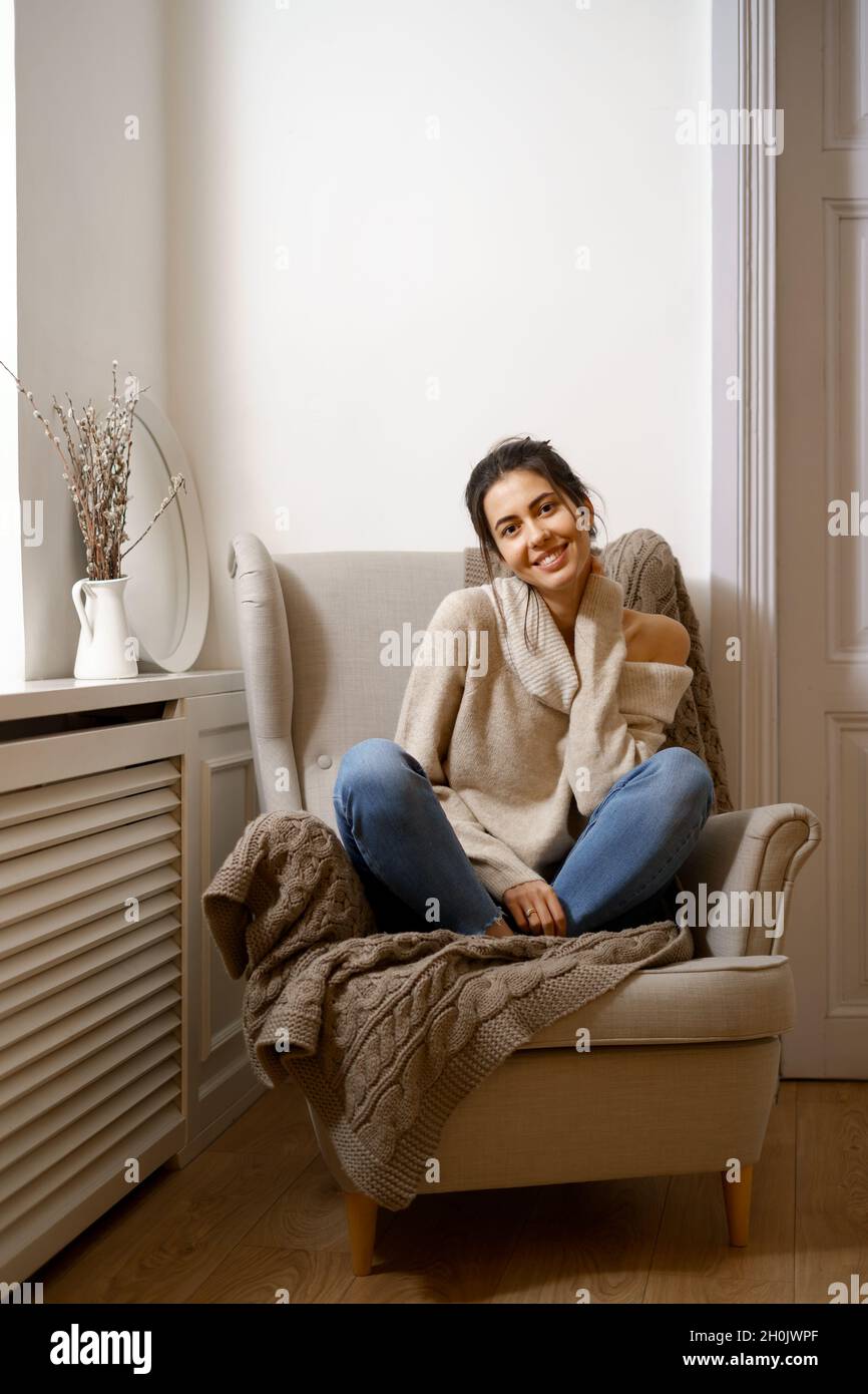 Señora sonriente con ropa elegante de moda está sentada en el sillón.  Sonriente, sentado en un ambiente relajante en casa, con un diseño  agradable. Comodidad y un tiempo agradable para las niñas