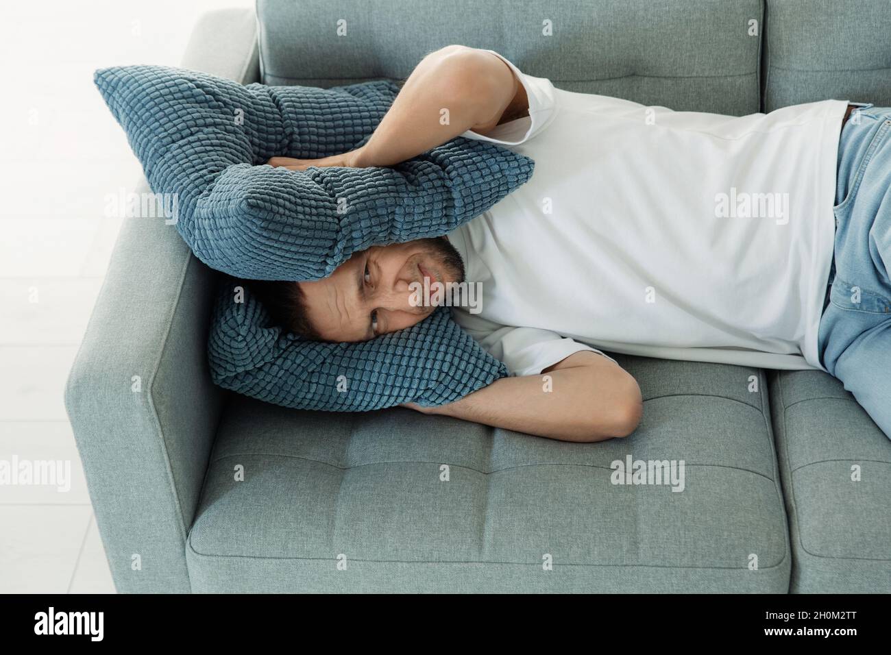 Hombre molesto cubrir las orejas con almohadas para evitar el ruido del vecino mientras siesta en el sofá tratar de dormir Foto de stock