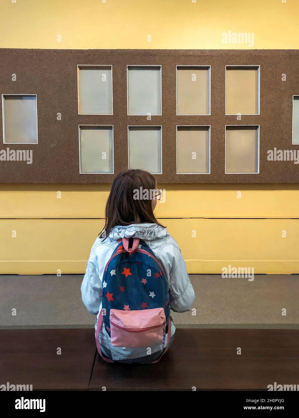 Vista posterior de la niña adolescente con mochila en el museo visitando la exposición, la colegiala sentada en el banco en la galería de arte y mirando con interés al objeto de arte Foto de stock