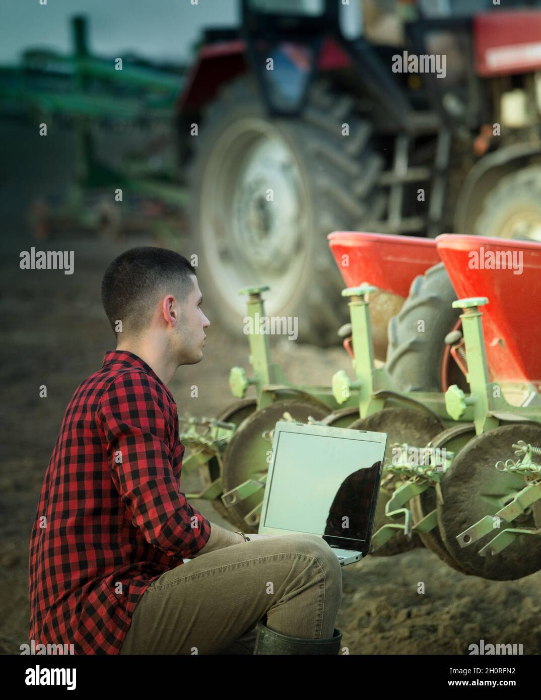 Joven agricultor con laptop en cuclillas en el campo en la parte delantera del tractor con equipo de siembra. Tecnología inteligente en la agricultura Foto de stock