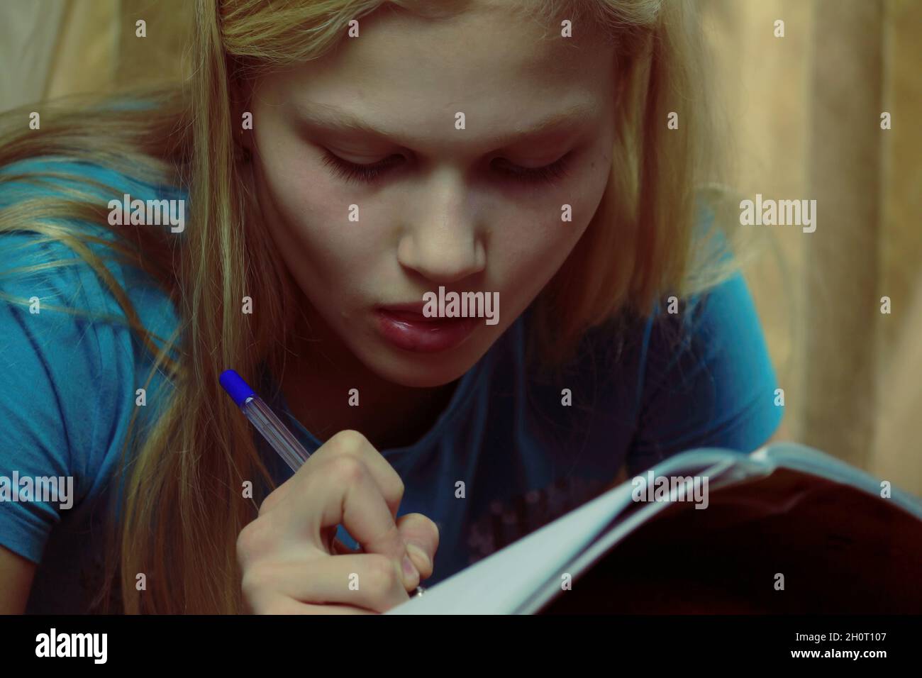 Una joven adolescente rubia está ocupada escribiendo en un cuaderno en el interior. Foto de stock