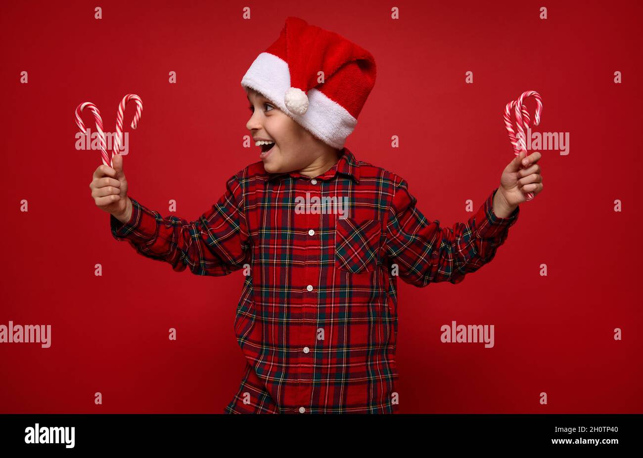 Hermoso niño con camisa de cuadros y sombrero de Santa Claus mirando a los  piruletas de Navidad, bastones de caramelo a rayas en sus manos, aislados  sobre rojo Fotografía de stock -