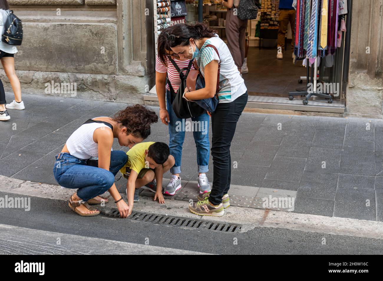 Mujer y niño tratando de recoger un objeto caído del desagüe de agua de lluvia en Roma, Italia Foto de stock