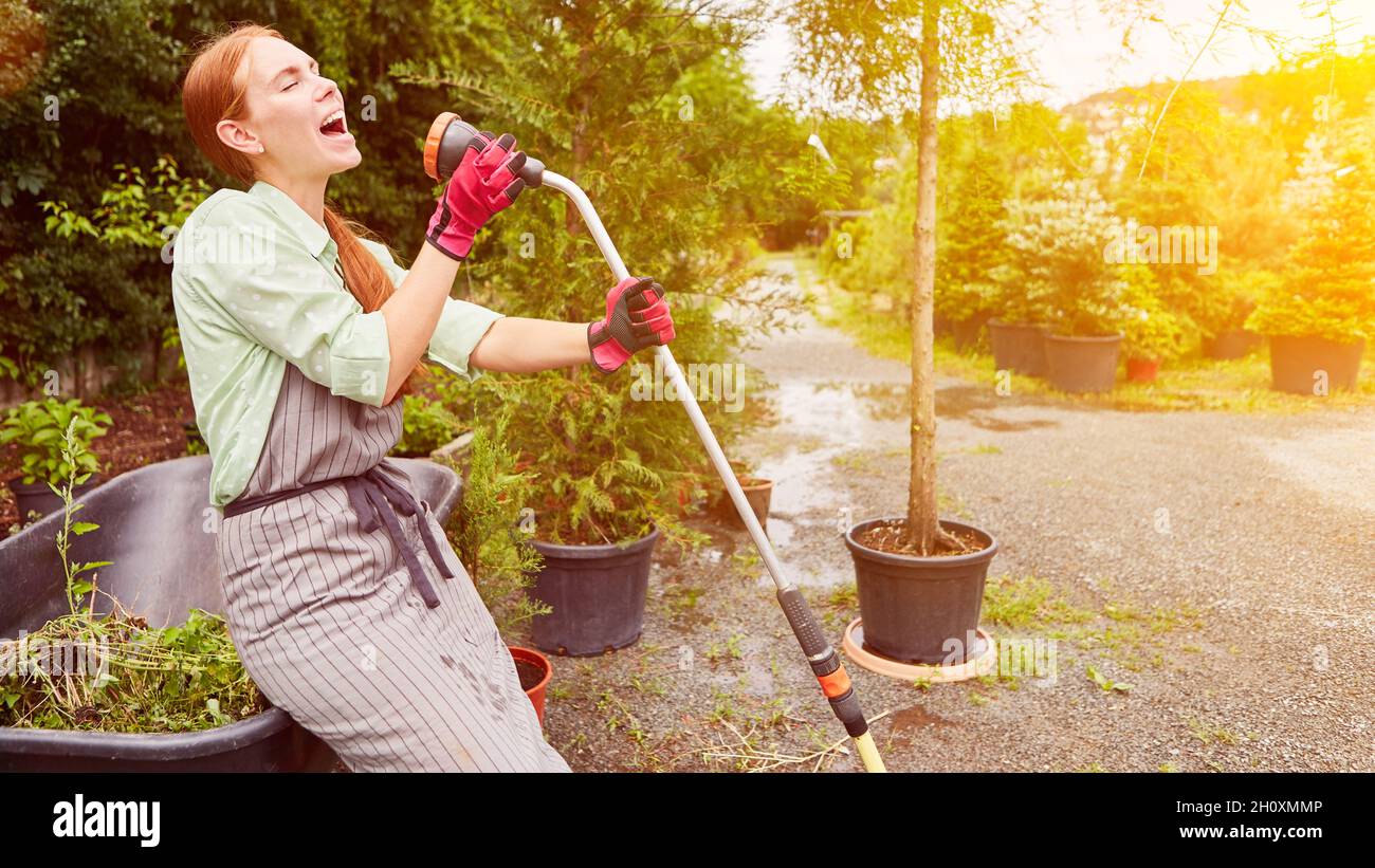 Mujer en el entrenamiento en jardinería tiene diversión en el trabajo y canta en una manguera de jardín Foto de stock
