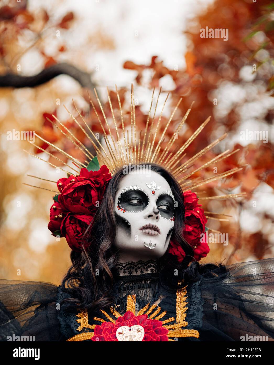 Retrato de una mujer joven con maquillaje de cráneo de azúcar y rosas rojas  vestidas con traje negro de muerte como Santa Muerte contra el fondo de  otoño se va Fotografía de