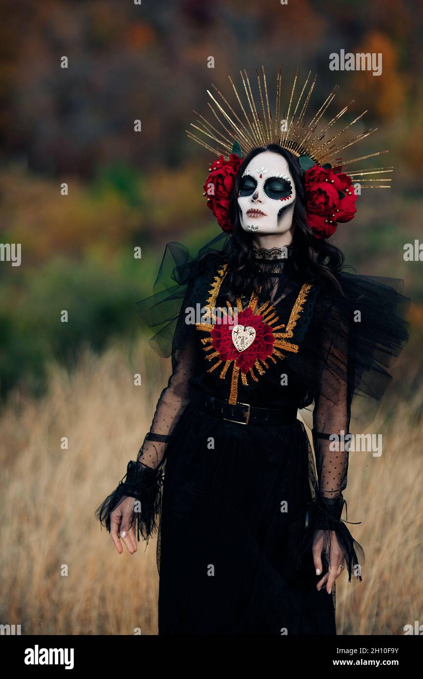 Mujer joven con maquillaje de cráneo de azúcar y rosas rojas vestidas con  traje negro de la muerte como Santa Muerte está contra el fondo del bosque  de otoño. Día del Fotografía