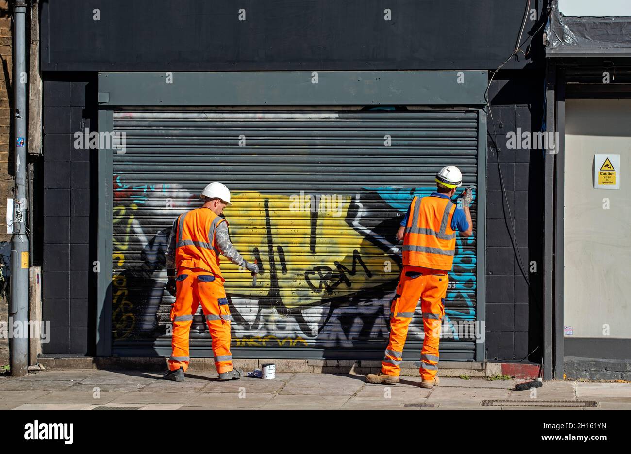 Dos hombres de trabajo con chalecos de alta visibilidad quitando graffiti de la tienda de la cortina pintando sobre él Londres Borough de Camden Inglaterra Reino Unido Foto de stock