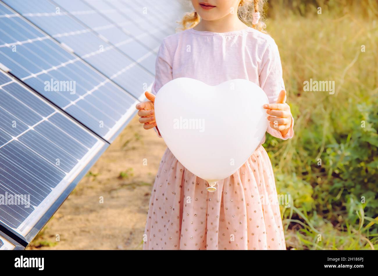 Niña irreconocible con globo blanco en forma de corazón vacío, de pie junto a paneles solares, granja solar en el soleado día de verano. Promover la energía verde Foto de stock