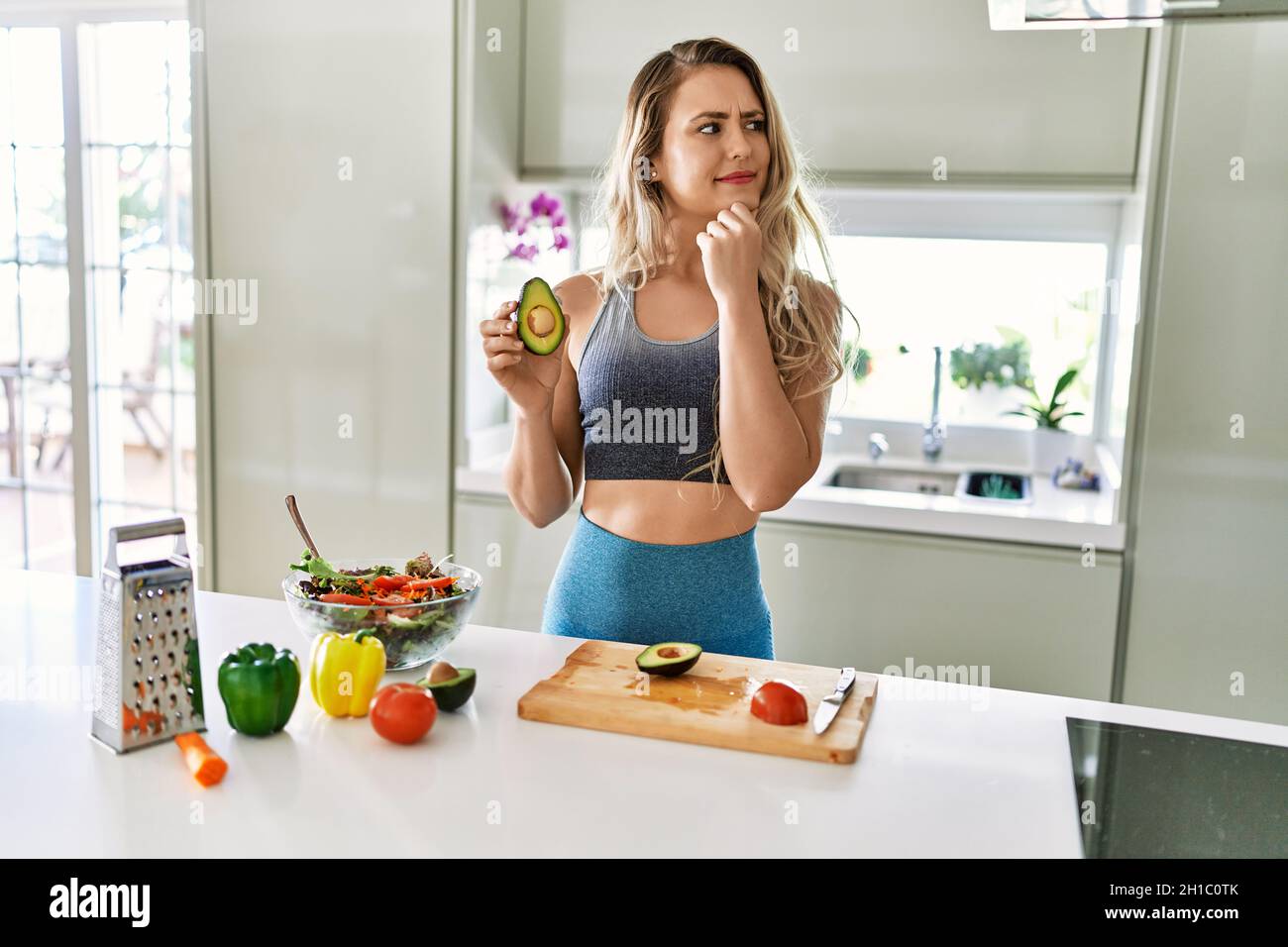 Joven mujer de fitness con ropa deportiva preparando ensalada saludable en la cocina seria cara la pregunta con la mano en barbilla, sin embargo Fotografía de stock -