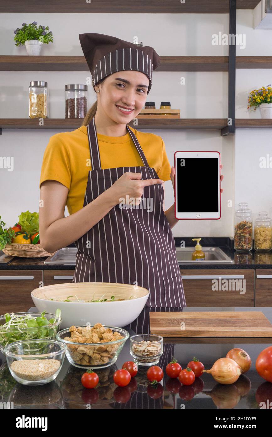 La ama de casa vestida con un delantal y un capuchón, sosteniendo una pantalla en blanco de la tableta de ordenador. Ambiente matutino en una cocina moderna. Foto de stock