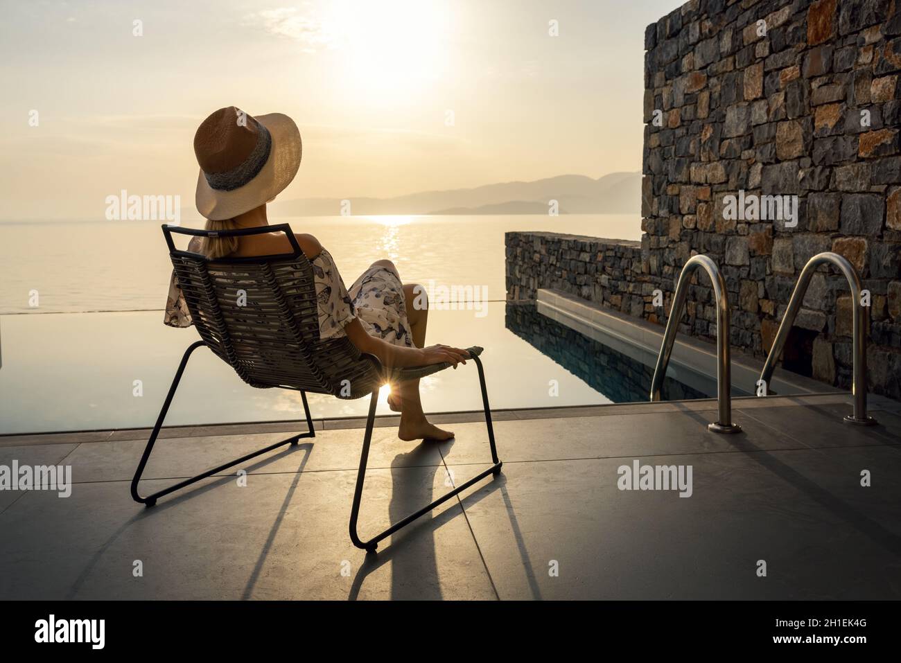 vacaciones relajantes - mujer sentada en la silla en la terraza frente a la piscina y disfrutando del amanecer en una villa de lujo Foto de stock