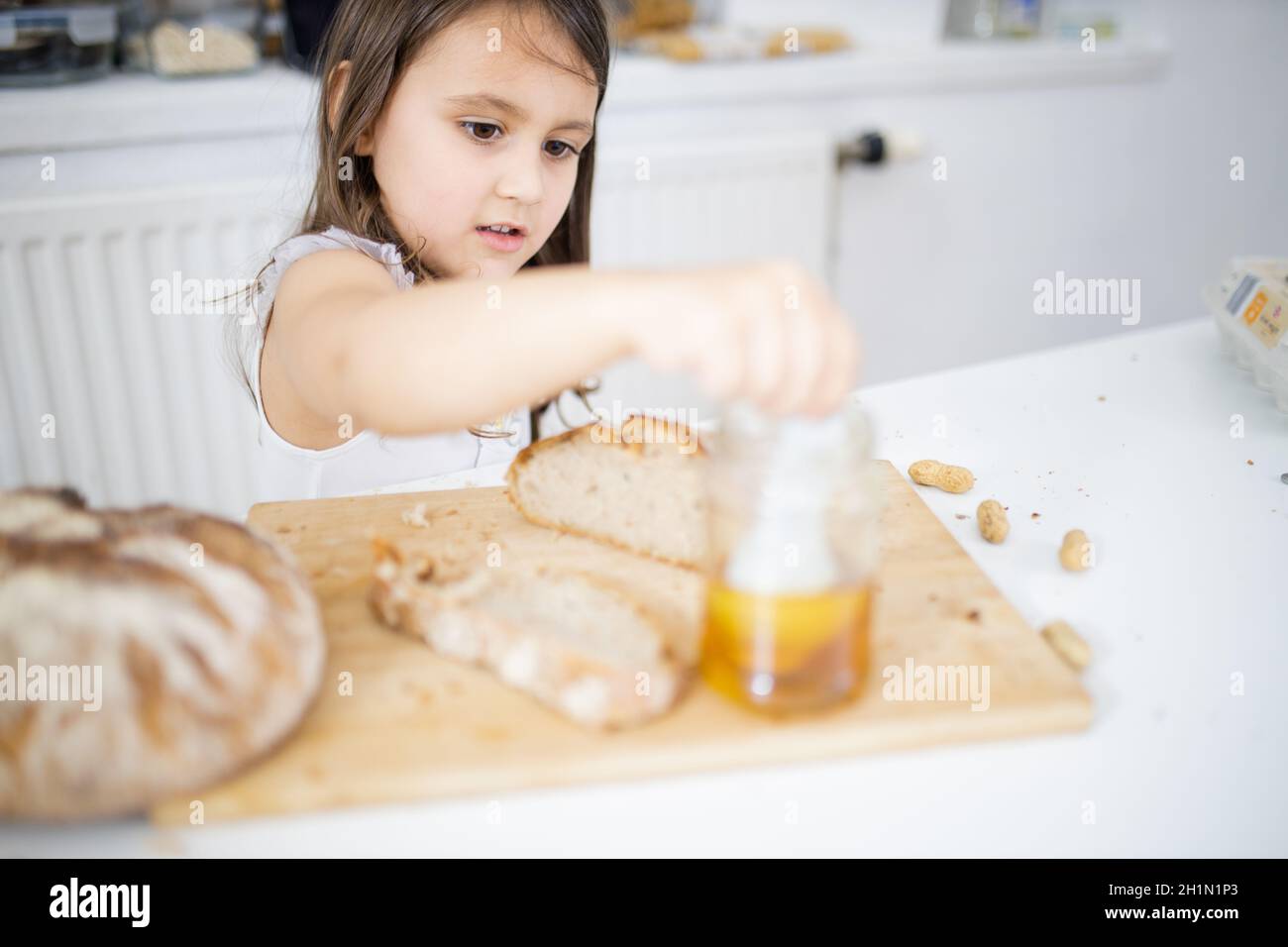Pequeña niña recogiendo miel del tarro junto a una rebanada de pan encima  de la tabla de madera. Niño pequeño en la mesa blanca pegando una cuchara  en el tarro de miel.