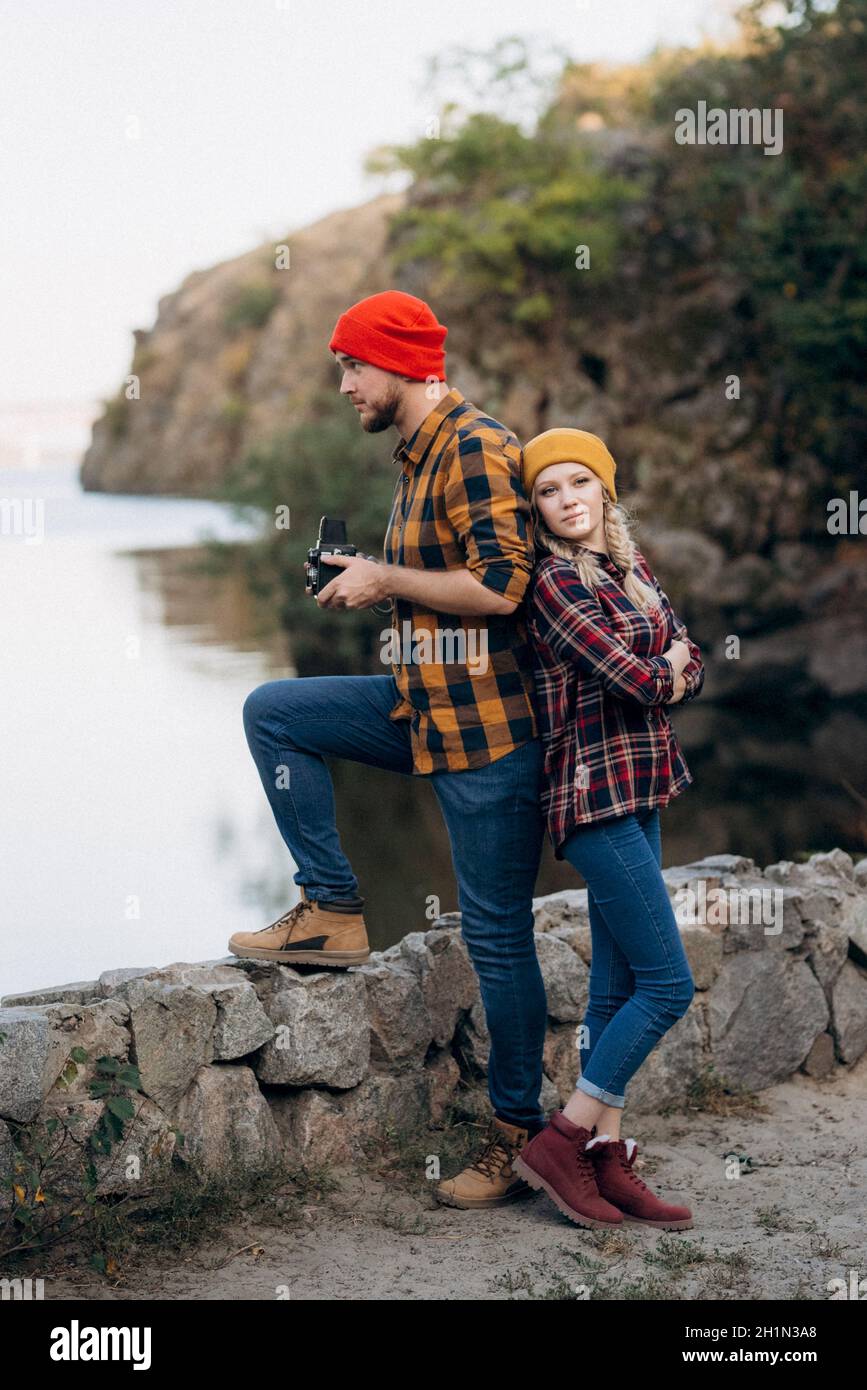 Un chico calvo con barba y una chica rubia sombreros brillantes en el fondo del río están tomando fotografías con una cámara antigua Foto de stock