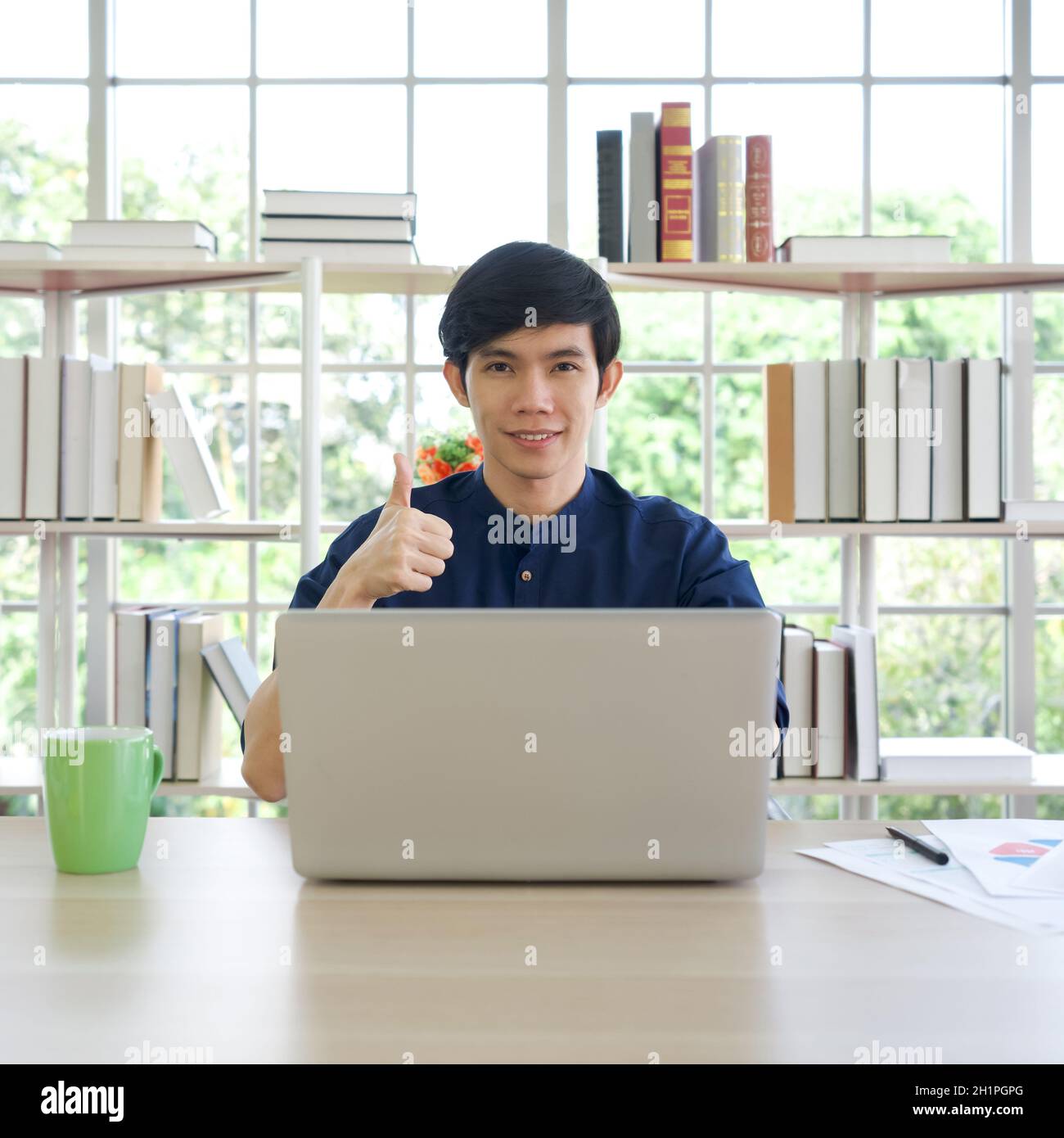Un joven asiático levanta el pulgar con una sonrisa detrás del ordenador portátil de la biblioteca. Hay taza de café, computadora, pluma, y papeles de tesis en la t Foto de stock