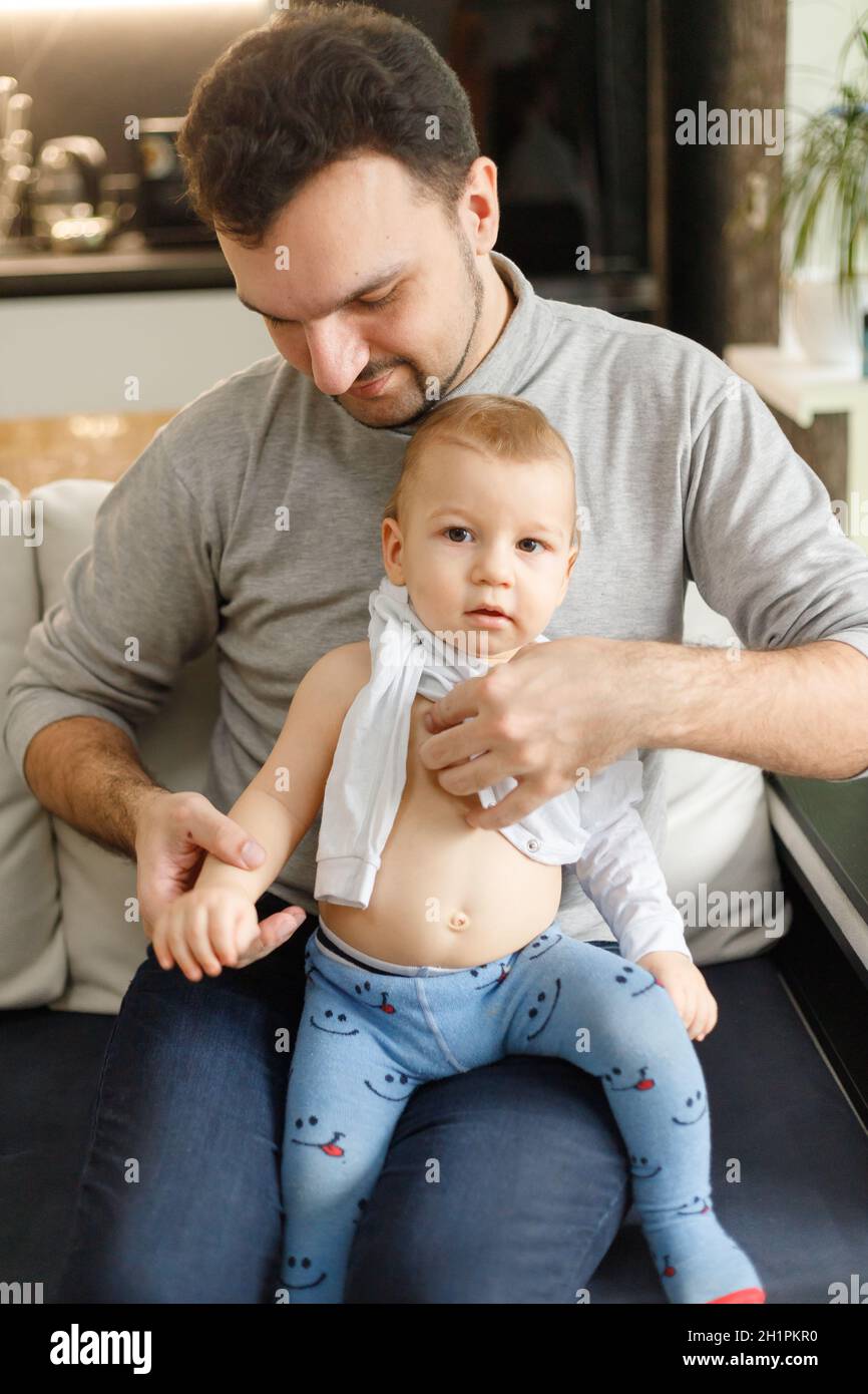 El padre viste al bebé en la sala de estar sentado en el sofá de la casa. Ropa  para bebés camisetas y deslizadores en el primer plano del bebé. Concepto  de cuidado