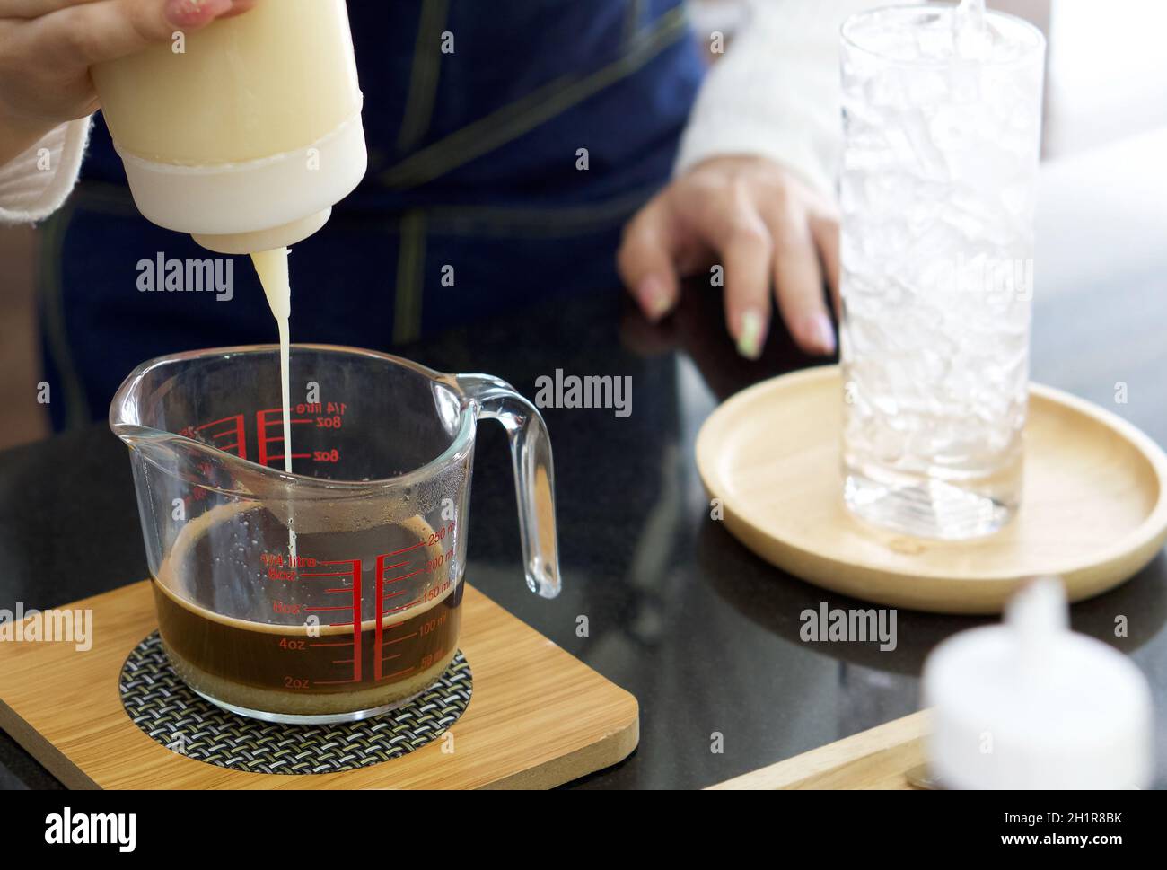 Un barista asiático exprime la leche condensada de una botella de plástico en una taza medidora de café. Ambiente matutino en una cafetería. Foto de stock
