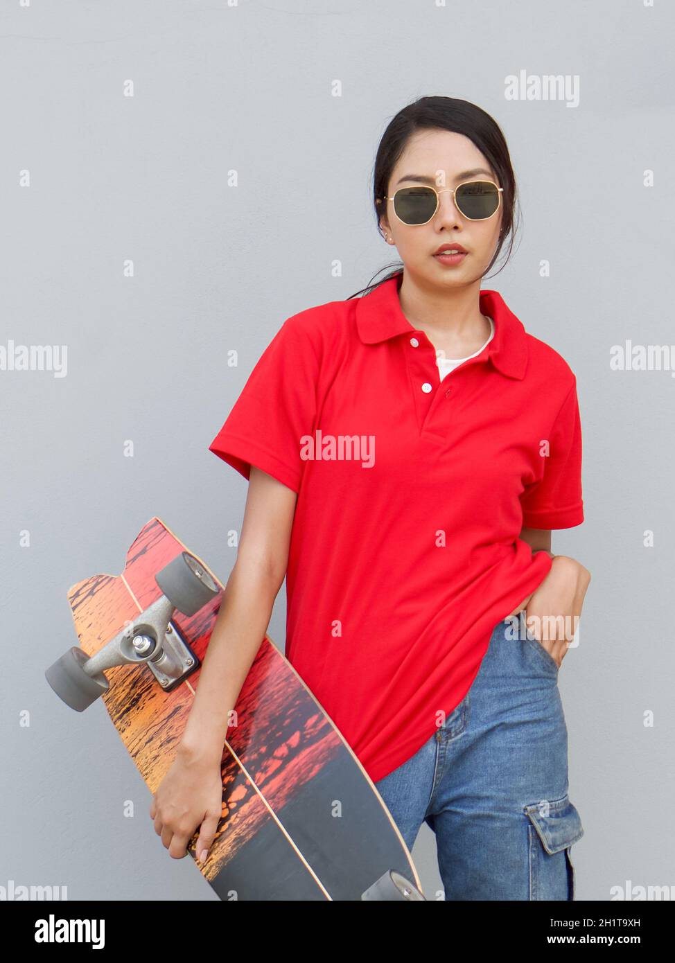Mujer asiática joven en camiseta roja con cuello sosteniendo la tabla de surf en posición de cadera. Mientras que ponga otra mano en el bolsillo de jean. Vertical sobre fondo gris Foto de stock