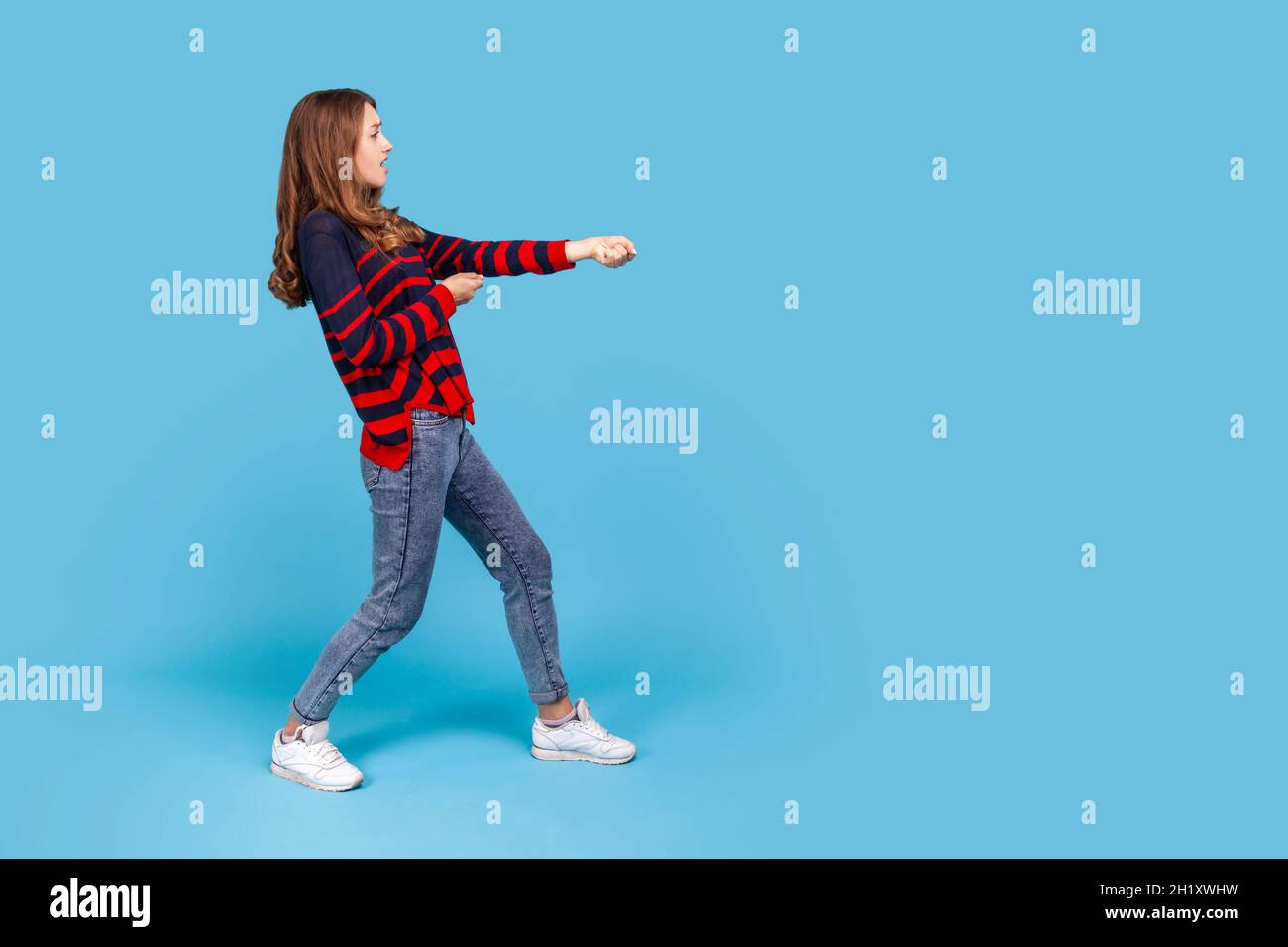 Retrato de perfil largo de mujer con suéter de rayas estilo sport, de pie en ataque o tirando de las manos gesto y mirando hacia delante. Estudio en interior grabado aislado sobre fondo azul. Foto de stock