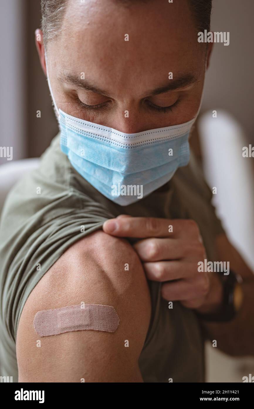 Hombre caucásico con mascarilla que muestra su hombro vacunado en casa Foto de stock