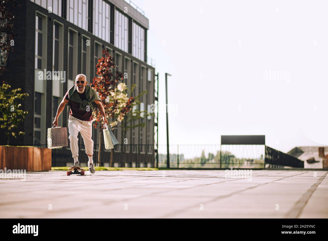 Hombre alegre con bolsas de compras para hacer skateboarding al aire libre Foto de stock