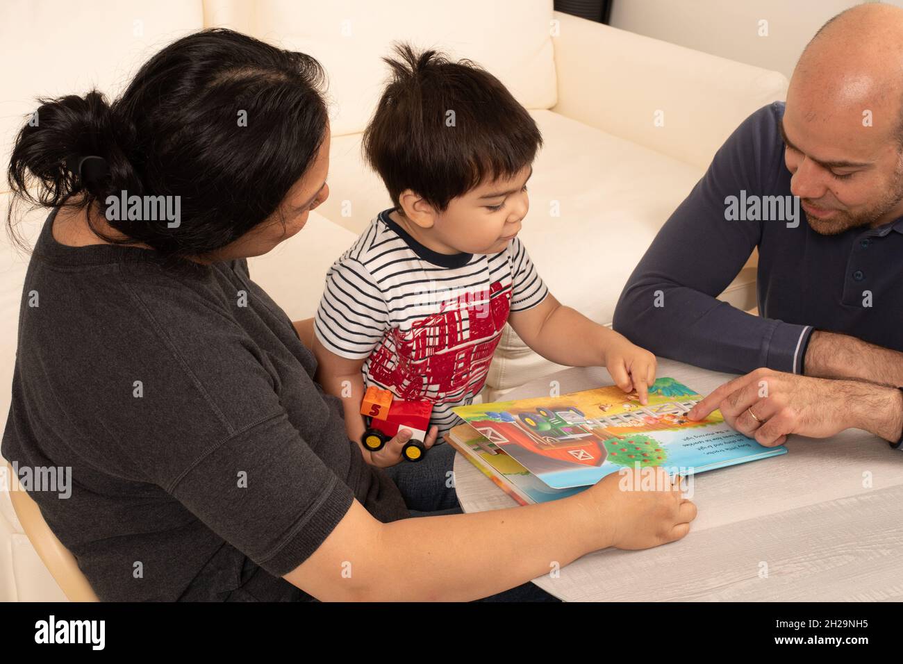 niño de 2 años con padres señalando y hablando, diciendo palabra mientras le leen el libro de fotos Foto de stock