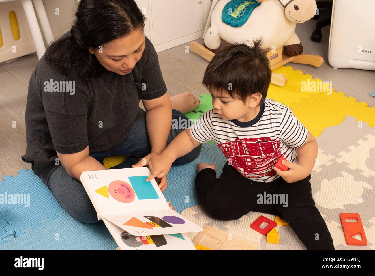 Niño de dos años que señala una forma cuadrada en el libro de la tabla madre está leyendo, mientras sostiene un bloque cuadrado en su mano Foto de stock