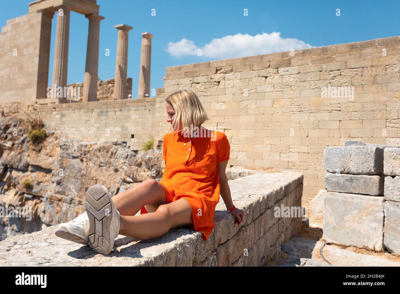 Una joven rubia está sentada y disfrutando de la vista de la costa cerca de la ciudad de Lindos, Grecia. Foto de stock