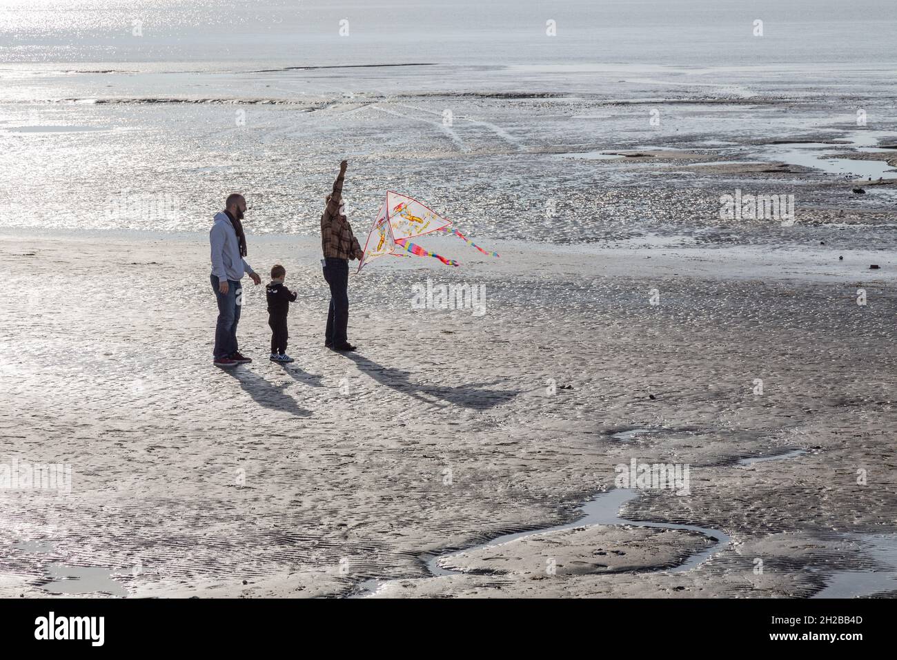 Tres personas, incluyendo un niño, manejando una cometa en una playa retroiluminada. Le Crotoy, Bahía de Somme, Francia Foto de stock