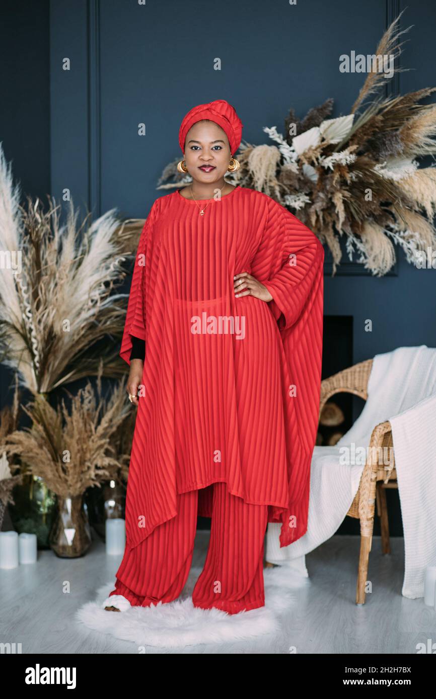 Joven modelo femenino africano atractivo en traje rojo con estilo y  pañuelo, posando en el estudio con decoraciones rústicas boho en el fondo.  Tiro completo de elegante dama africana Fotografía de stock -