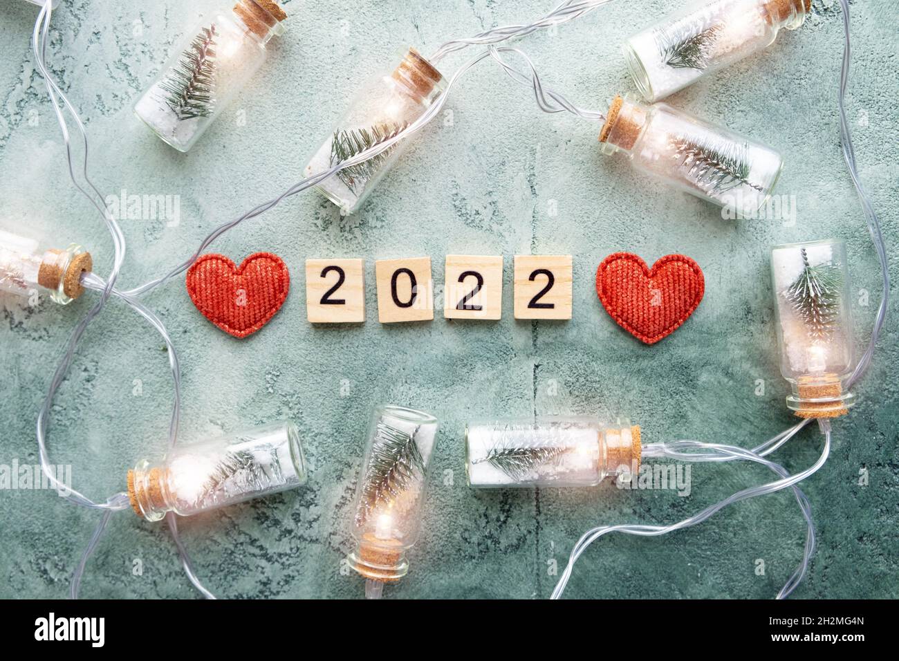 Número 2022 en cuadrados de madera y una brillante guirnalda de tarros de vidrio con árboles de Navidad y nieve en el interior y dos corazones sobre un fondo verde texturizado Foto de stock