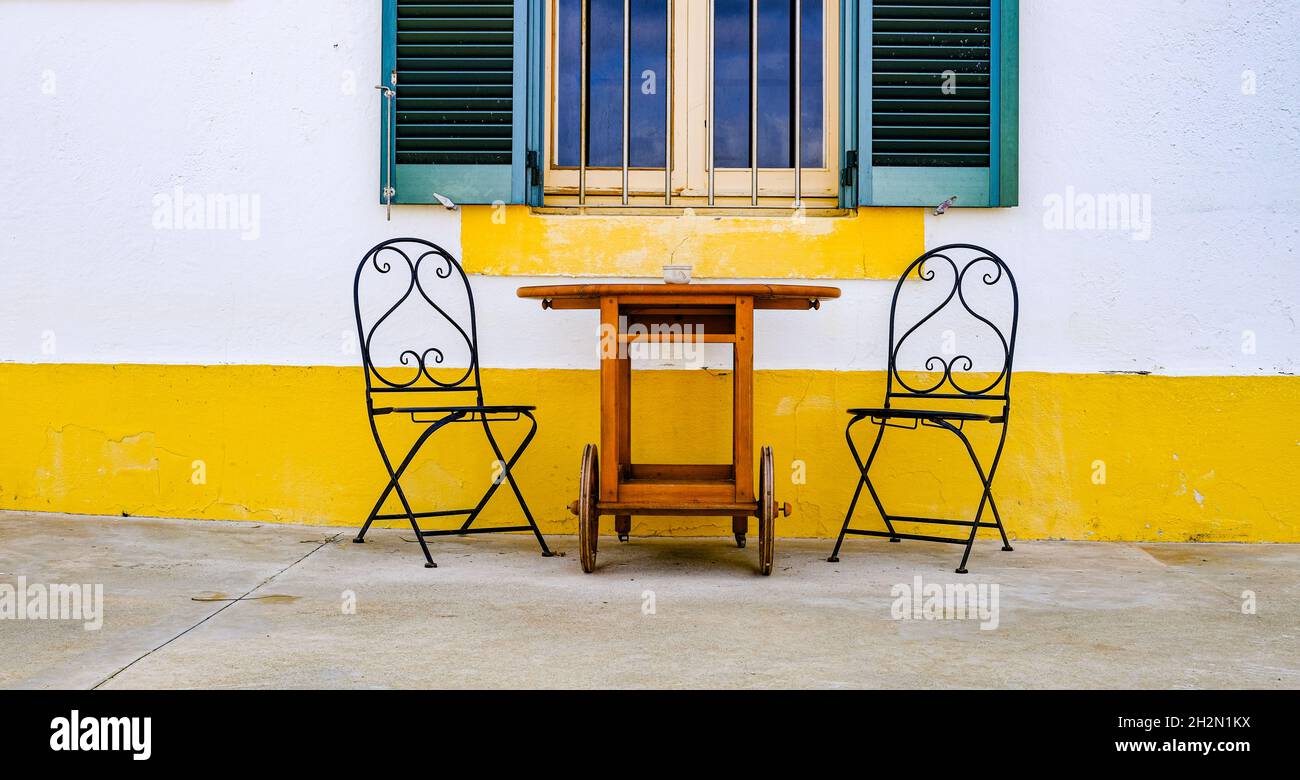 dos sillas de hierro y una mesa de madera con una ventana y una pared amarilla en la parte inferior. Foto de stock