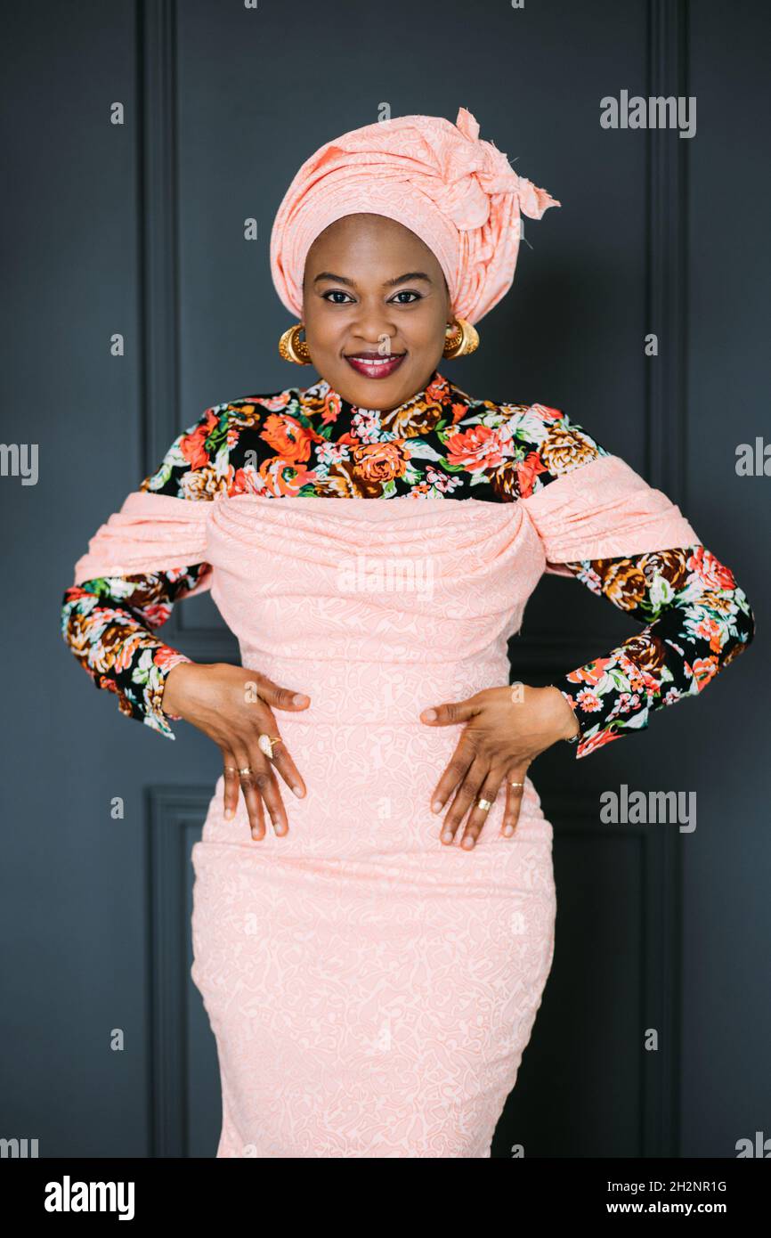 Retrato de encantadora mujer africana con elegante vestido rosa y turbante  en la cabeza, sonriendo en la cámara con los brazos en la cintura, posando  sobre fondo de pared azul oscuro Fotografía