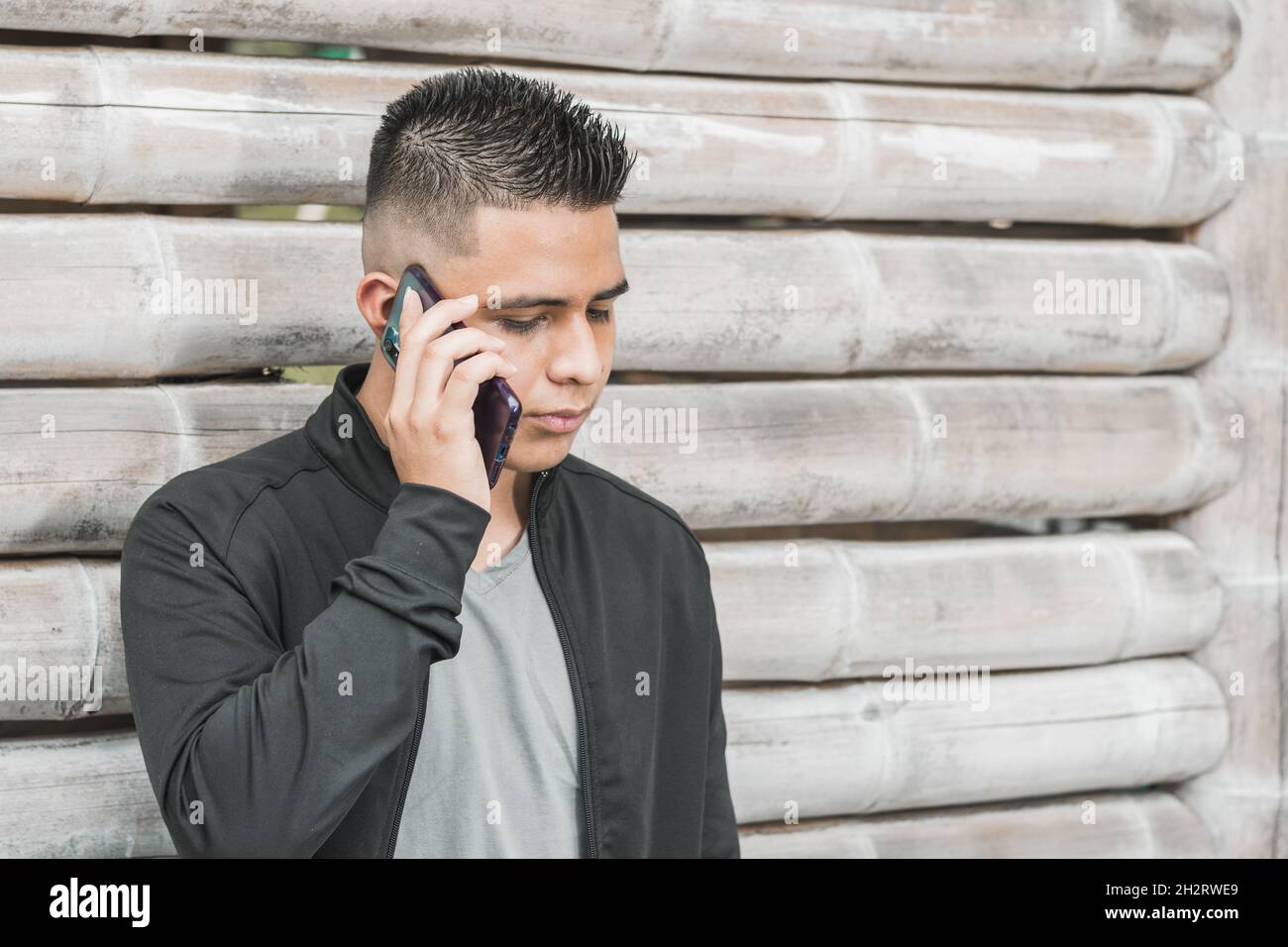 joven estudiante universitario latino hablando en su teléfono celular con una cara seria, apoyándose en una choza guadua, empresario o influencer con su teléfono celular Foto de stock
