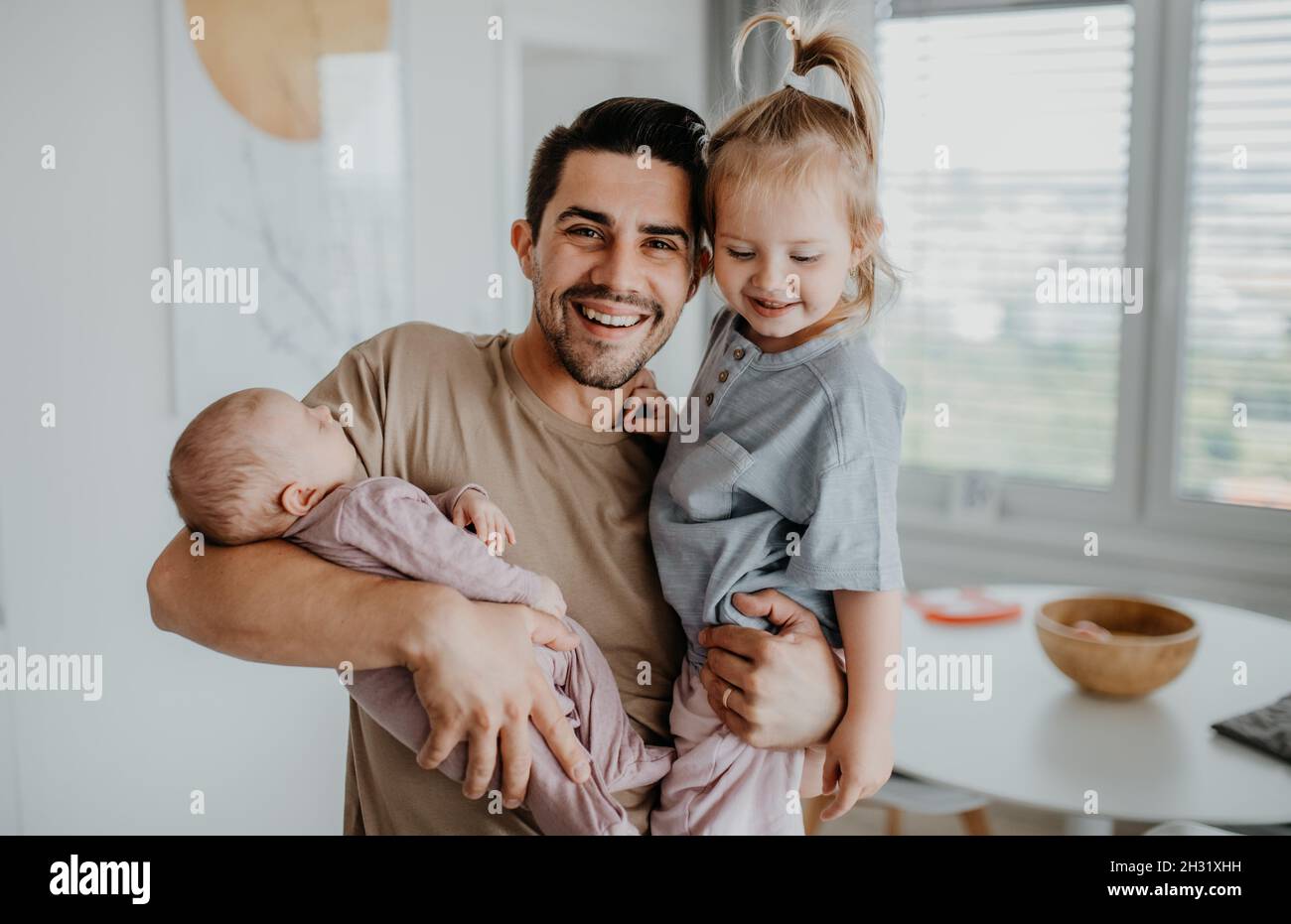 Feliz joven cuidando a su bebé recién nacido y a su hija pequeña en casa, permiso de paternidad Foto de stock