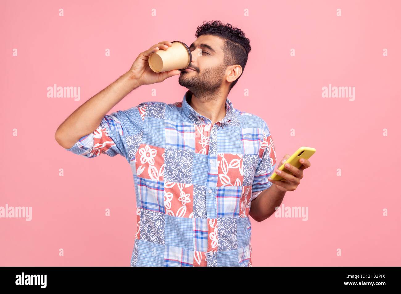 Retrato de un joven adulto atractivo en camisa de estilo informal de pie y sosteniendo una taza y un teléfono desechables, bebiendo y disfrutando de una bebida caliente. Estudio en interior grabado aislado sobre fondo rosa. Foto de stock