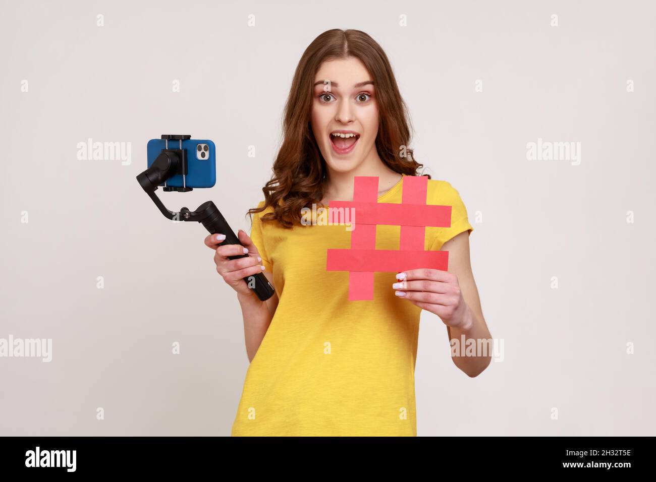 Una adolescente emocionada con el teléfono en el estabilizador lleva al videoblog, mirando la cámara con una expresión de alegría, mostrando el símbolo rojo de etiqueta hashtag. Estudio en interior grabado aislado sobre fondo gris. Foto de stock